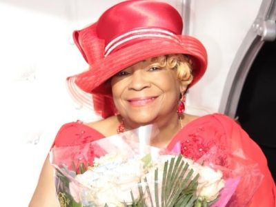 Beverly Tate is wearing a red dress, red hat and holding a flower bouquet.